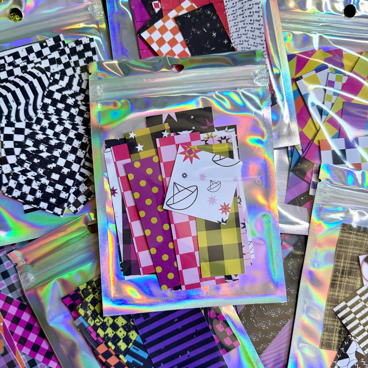 Several holographic zip-top pouches filled with colorful patterned sticker paper sheets arranged on a textured green surface. The papers feature various designs including stripes, polka dots, checkers, and abstract prints in pink, purple, yellow, black, white, and blue.