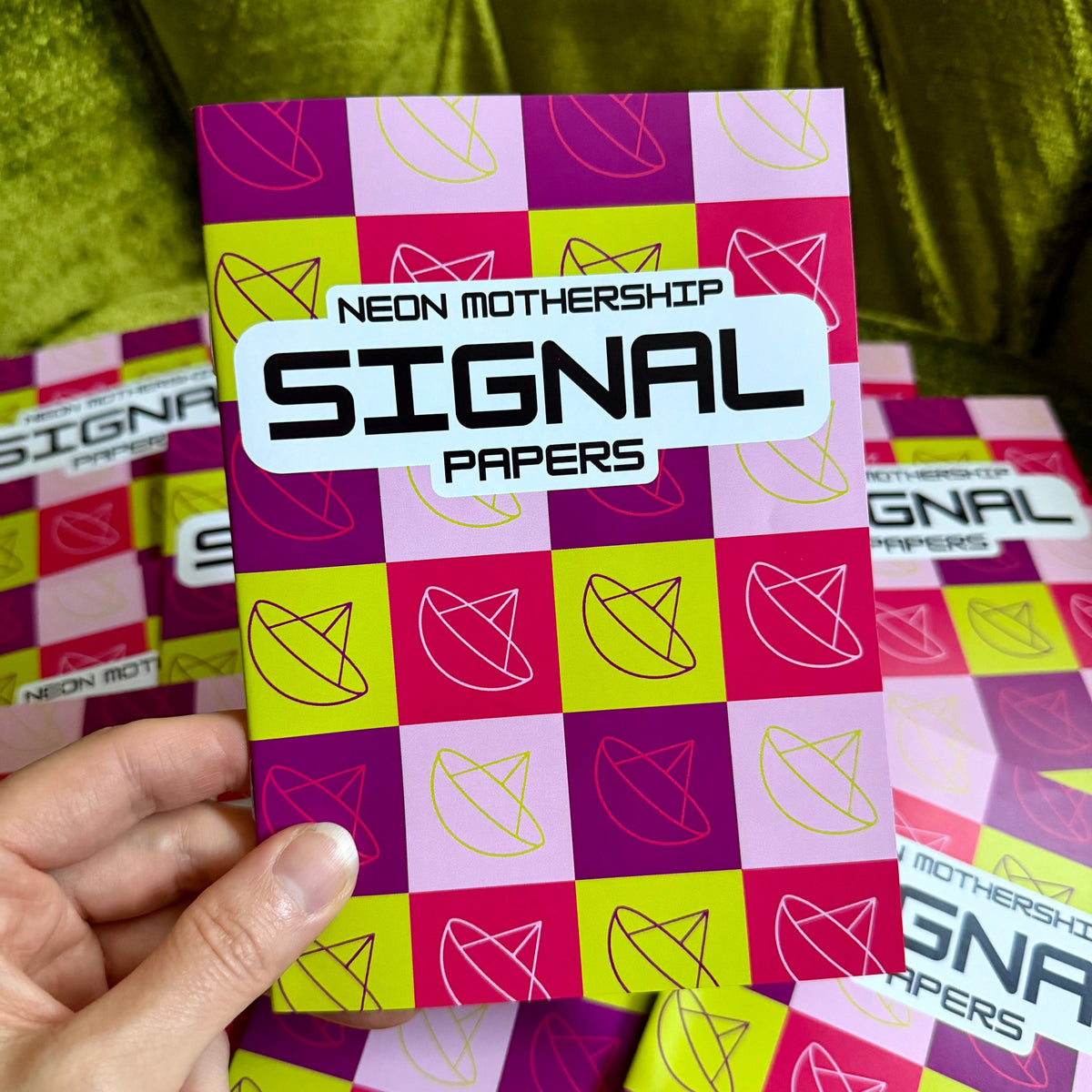 A hand holds a pack of 'Signal Papers' by Neon Mothership, featuring a vibrant neon pink, purple, and lime green checkered pattern with minimalist line drawings. More packs are scattered in the background on a shiny olive green velvet surface.