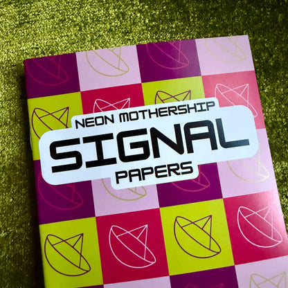 A notebook or paper pack titled 'NEON MOTHERSHIP SIGNAL PAPERS' with a colorful neon checkered cover in pink, purple, and yellow, photographed on a textured green surface.