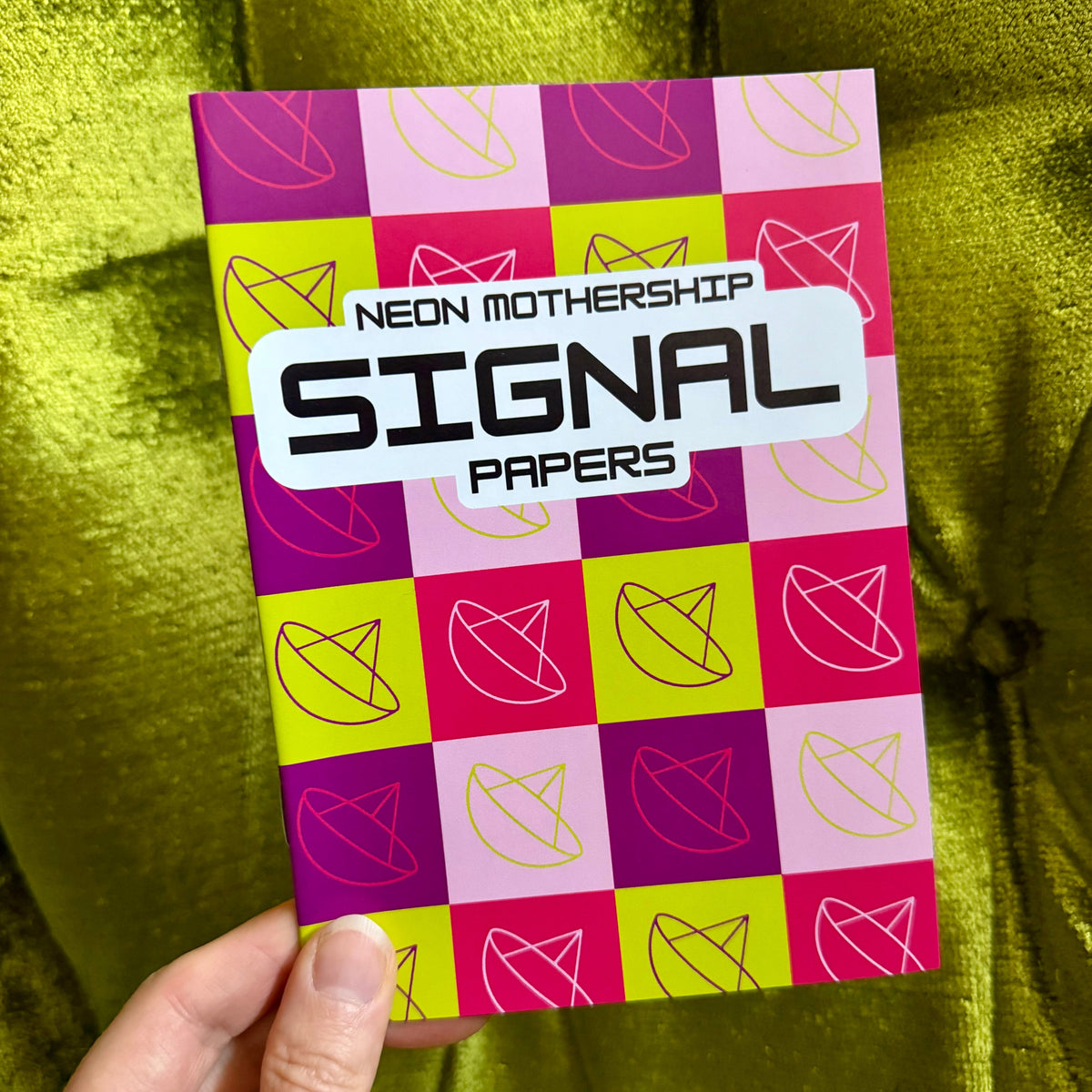 A hand holds a colorful card or packet labeled 'NEON MOTHERSHIP SIGNAL PAPERS' against a shiny olive-green fabric background. The product has a pink, purple, and yellow checkered pattern with geometric line drawings.