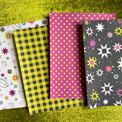 Four patterned paper sticker sheets arranged overlapping on a textured metallic gold background. The patterns include white with pink and black star motifs, yellow and black gingham, solid pink with yellow polka dots, and dark gray with pink, white, and yellow starburst motifs.