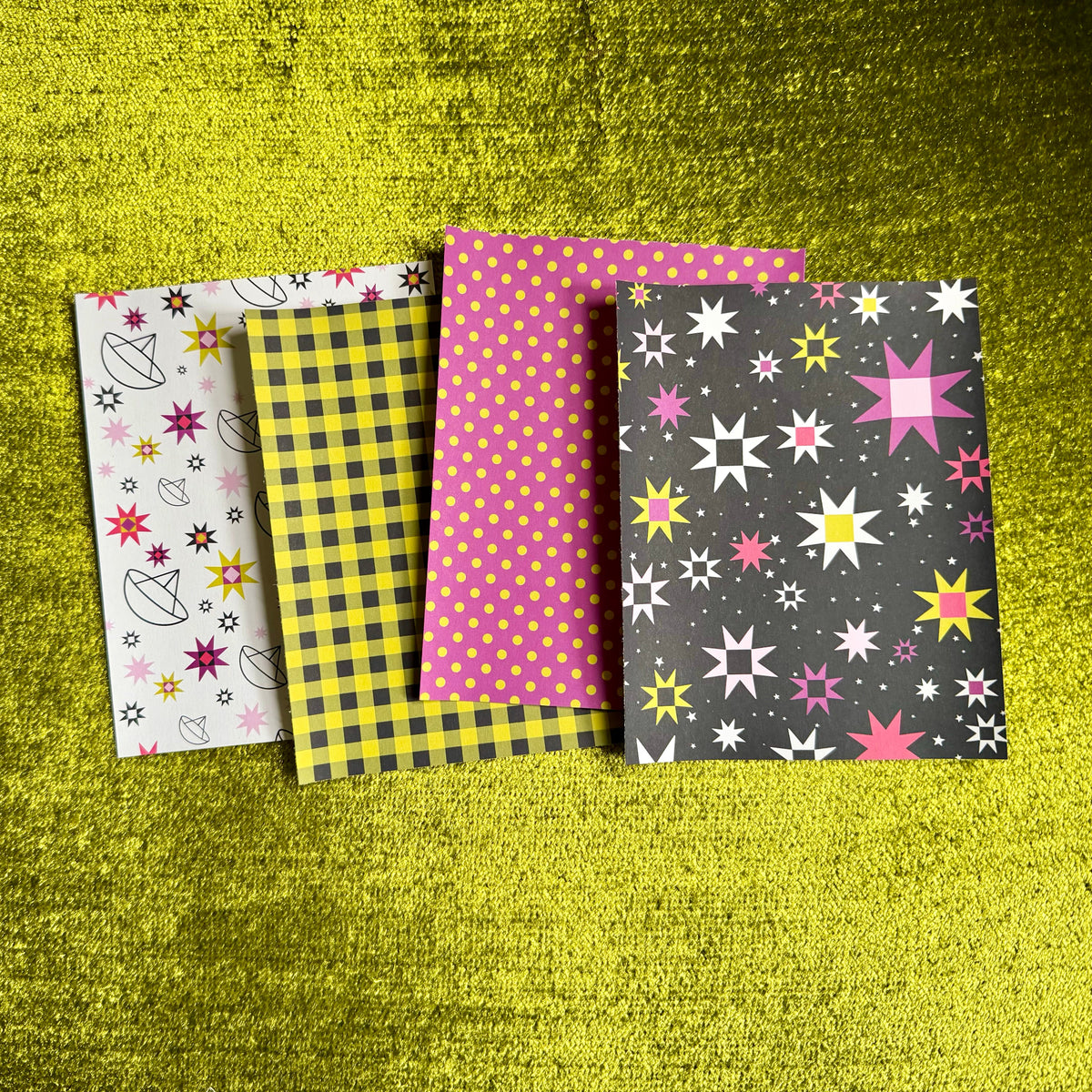 Four patterned sticker paper sheets arranged on a textured bright yellow-green surface. The designs include a white sheet with colorful star and flower motifs, a yellow-and-black gingham pattern, a pink-and-yellow polka dot pattern, and a dark gray sheet with colorful starbursts.