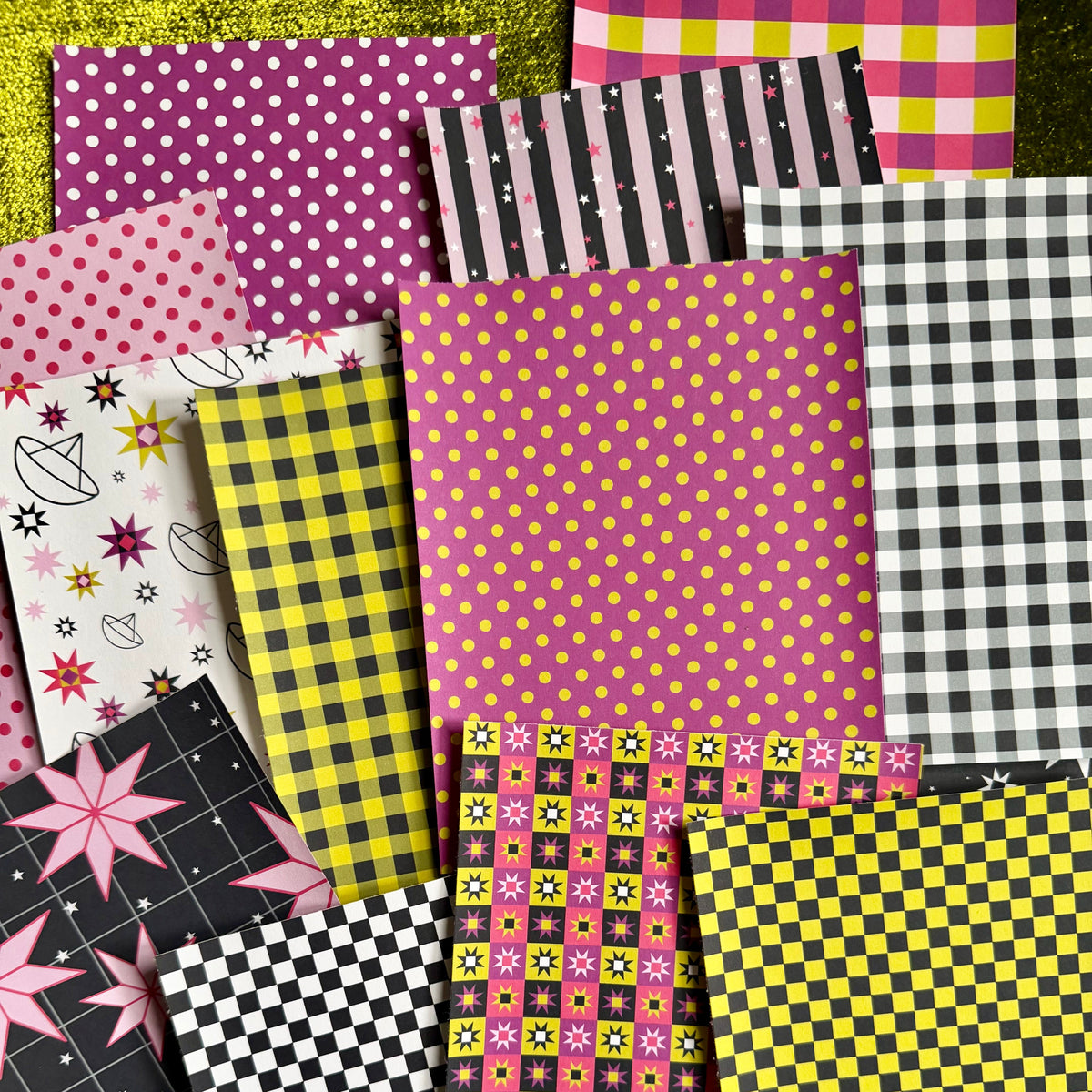 A flat-lay arrangement of multiple patterned sticker paper sheets in pink, yellow, black, white, and gray, including polka dots, stripes, gingham, stars, and checkerboard designs, set against a textured green background.