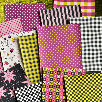 A flat-lay arrangement of multiple patterned sticker paper sheets in pink, yellow, black, white, and gray, including polka dots, stripes, gingham, stars, and checkerboard designs, set against a textured green background.