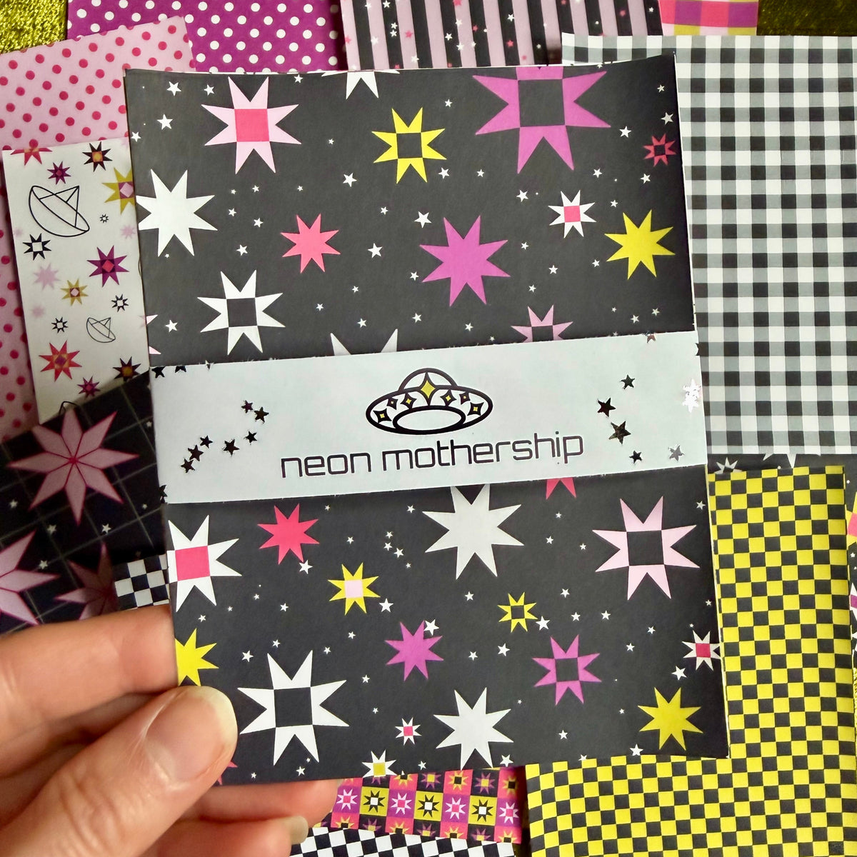 A hand holds a patterned sticker sheet set featuring a black background with neon pink, yellow, and white starburst designs. The set has a white band across the middle with the text 'neon mothership' and a small UFO graphic.