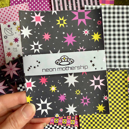 A hand holds a patterned sticker sheet set featuring a black background with neon pink, yellow, and white starburst designs. The set has a white band across the middle with the text 'neon mothership' and a small UFO graphic.