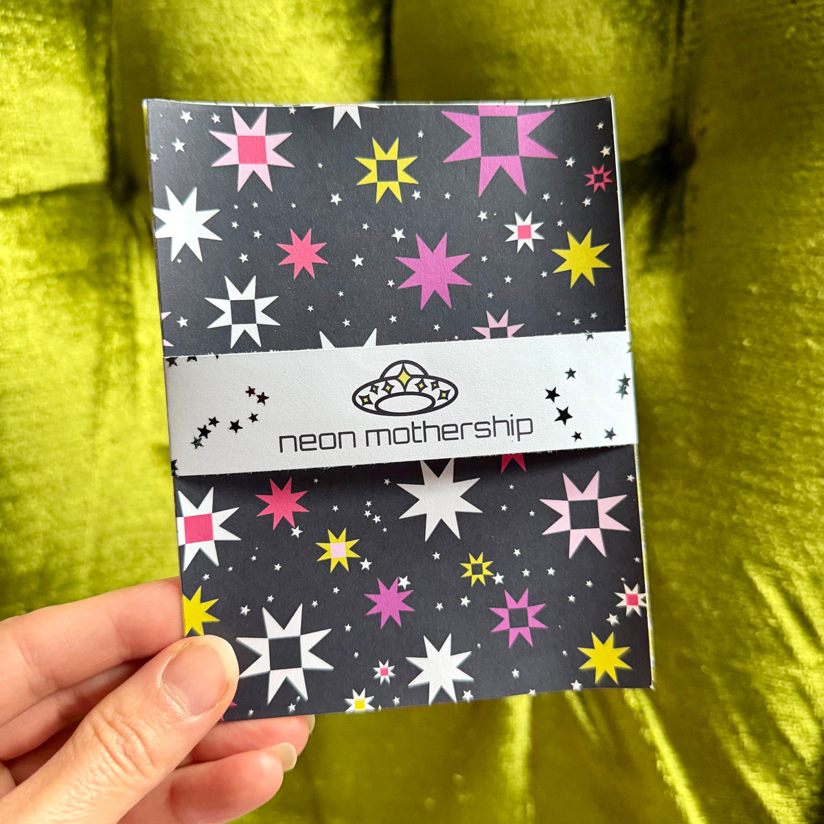 A hand holding a small sticker sheet set with a dark gray background and colorful starburst pattern in pink, yellow, and white. The center band reads 'neon mothership' with a small alien logo.