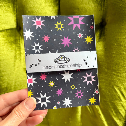 A hand holding a small sticker sheet set with a dark gray background and colorful starburst pattern in pink, yellow, and white. The center band reads 'neon mothership' with a small alien logo.