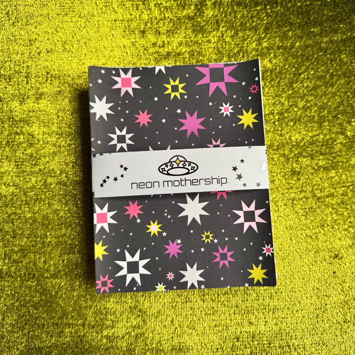 A bundle of sticker paper with a colorful star pattern, wrapped with a white band reading 'neon mothership' and a small alien logo, placed on a bright green textured surface.