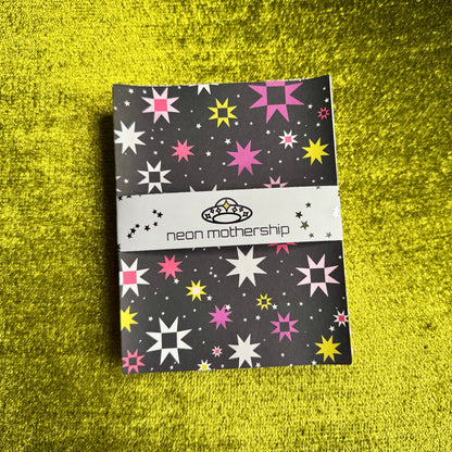 A bundle of sticker paper with a colorful star pattern, wrapped with a white band reading 'neon mothership' and a small alien logo, placed on a bright green textured surface.