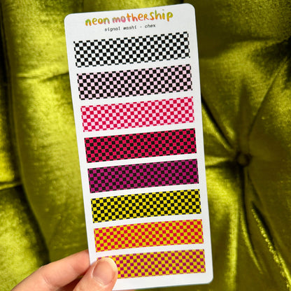 A hand holds a white sticker sheet displaying eight checkered pattern strips in neon shades of black, pink, red, purple, yellow, orange, and brown, set against a shiny olive green velvet background.
