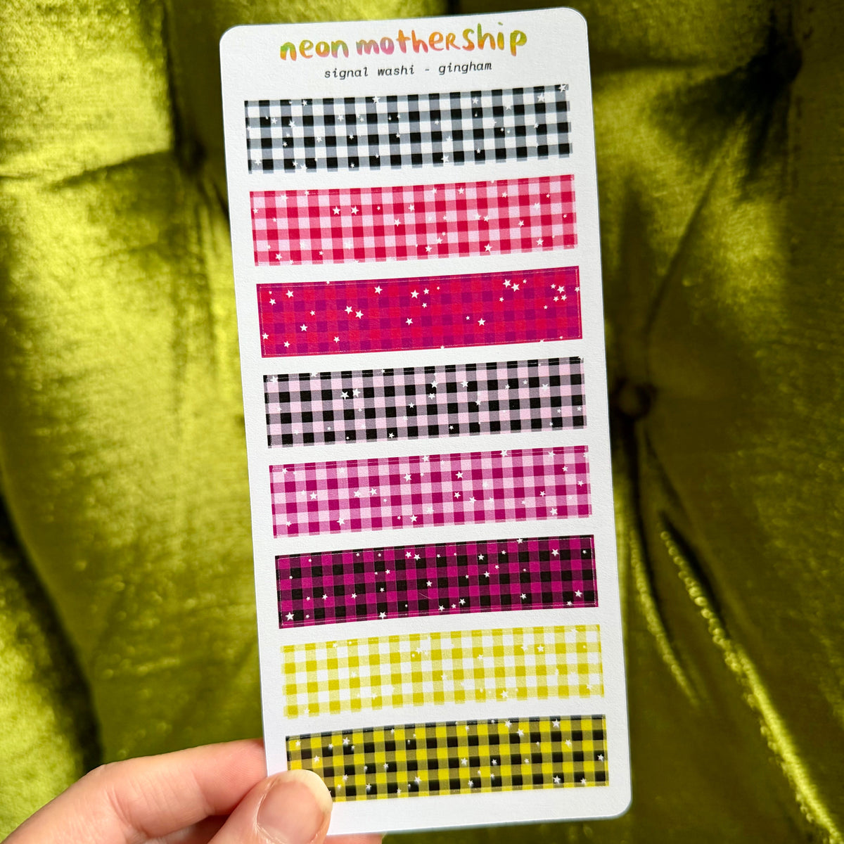 A hand holds a white card displaying neon mothership gingham patterns in various colors, including pink, yellow, black, and purple, set against a shiny olive green velvet background.