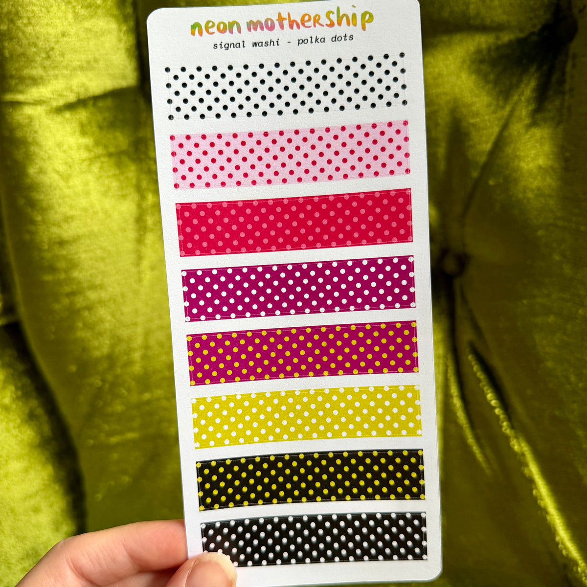A hand holds a white sticker sheet displaying eight horizontal strips of neon-colored polka dot washi tape in shades of black, pink, red, purple, yellow, and orange, set against a shiny olive green velvet background.