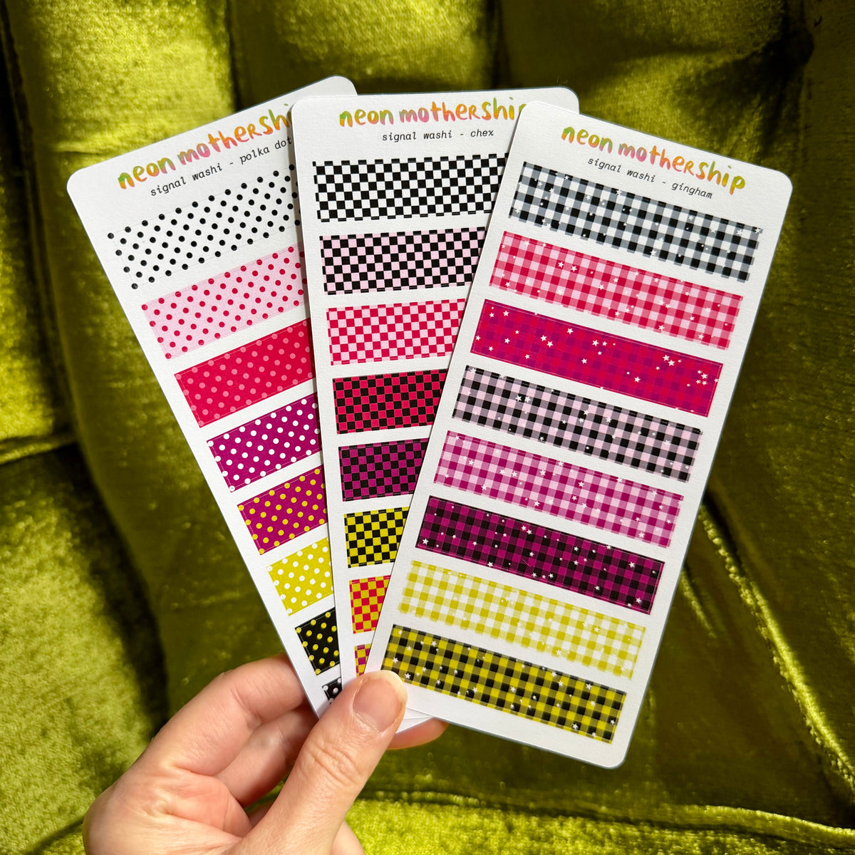 A hand holds three patterned planner sticker sheets against a shiny olive-green velvet background. The sheets feature neon pink, red, yellow, black, and white patterns including polka dots, gingham, and checkered designs.