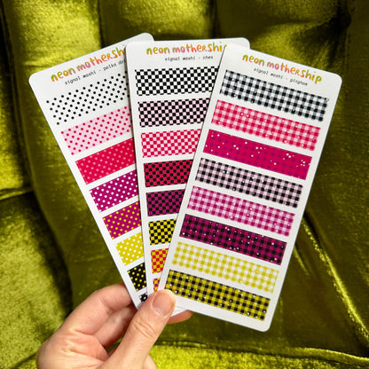 A hand holds three patterned planner sticker sheets against a shiny olive-green velvet background. The sheets feature neon pink, red, yellow, black, and white patterns including polka dots, gingham, and checkered designs.