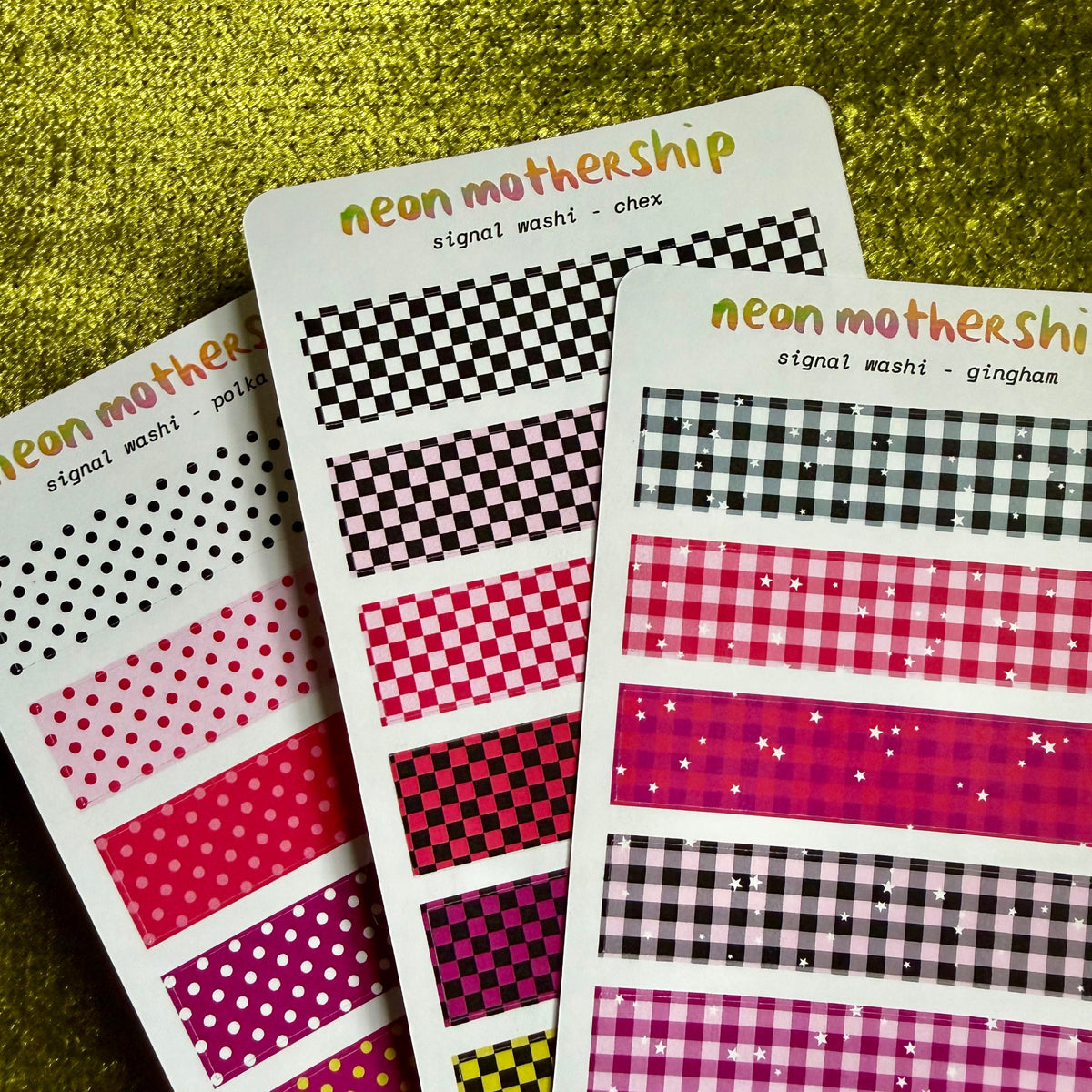 Three sheets of washi tape with different pattern designs, including checkers, gingham, polka dots, and glitter stripes, in vibrant pink, red, yellow, and black. The sheets are branded 'neon mothership' and displayed on a textured green gold background.