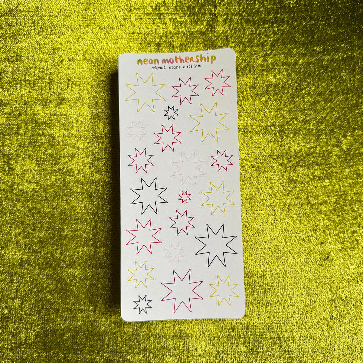 A sheet of neon star stickers on a textured yellow background. The stickers feature various star designs outlined in pink, yellow, and black, with some filled-in stars in pink and yellow.