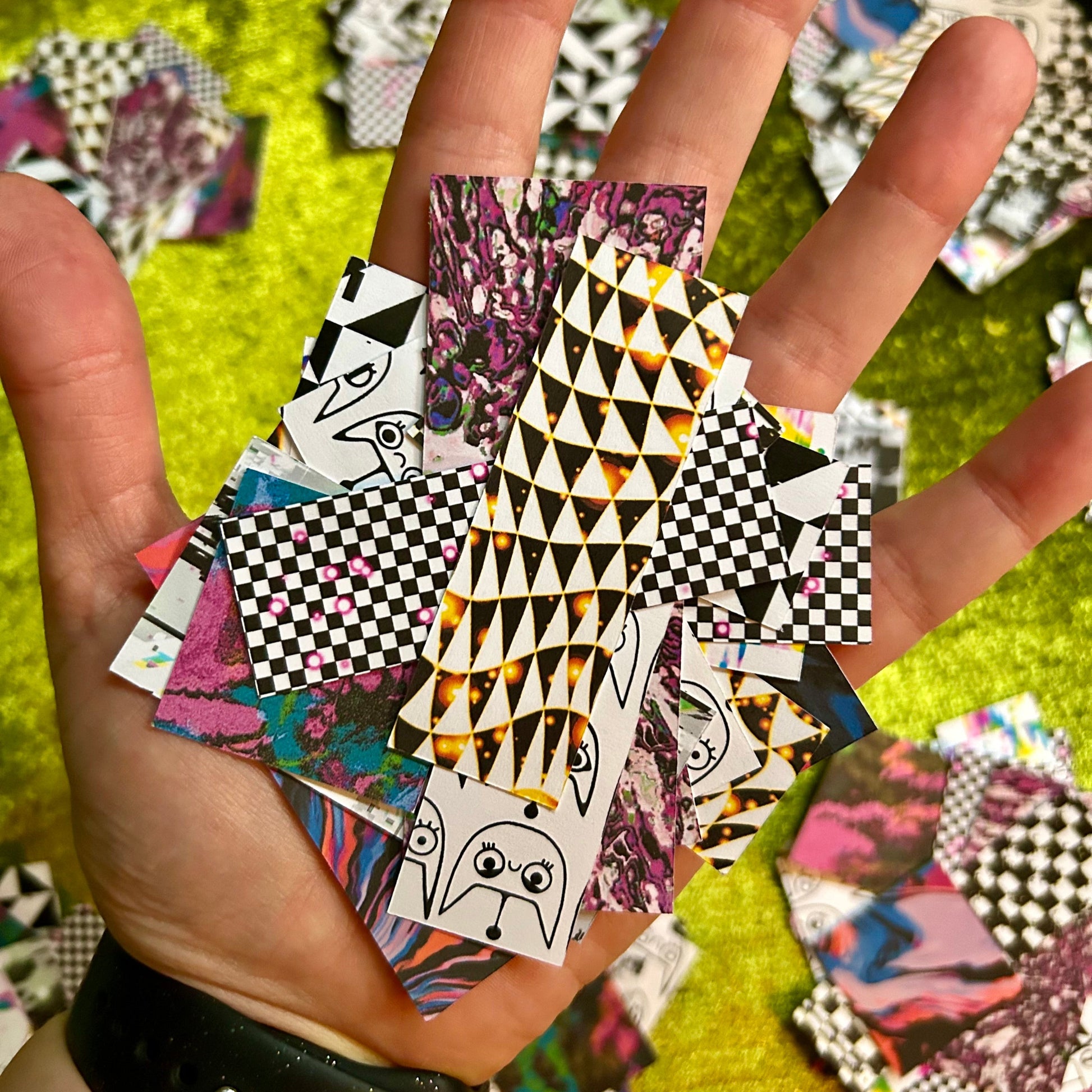 Hand holding a collection of small, colorful patterned sticker scraps against a green fabric background.