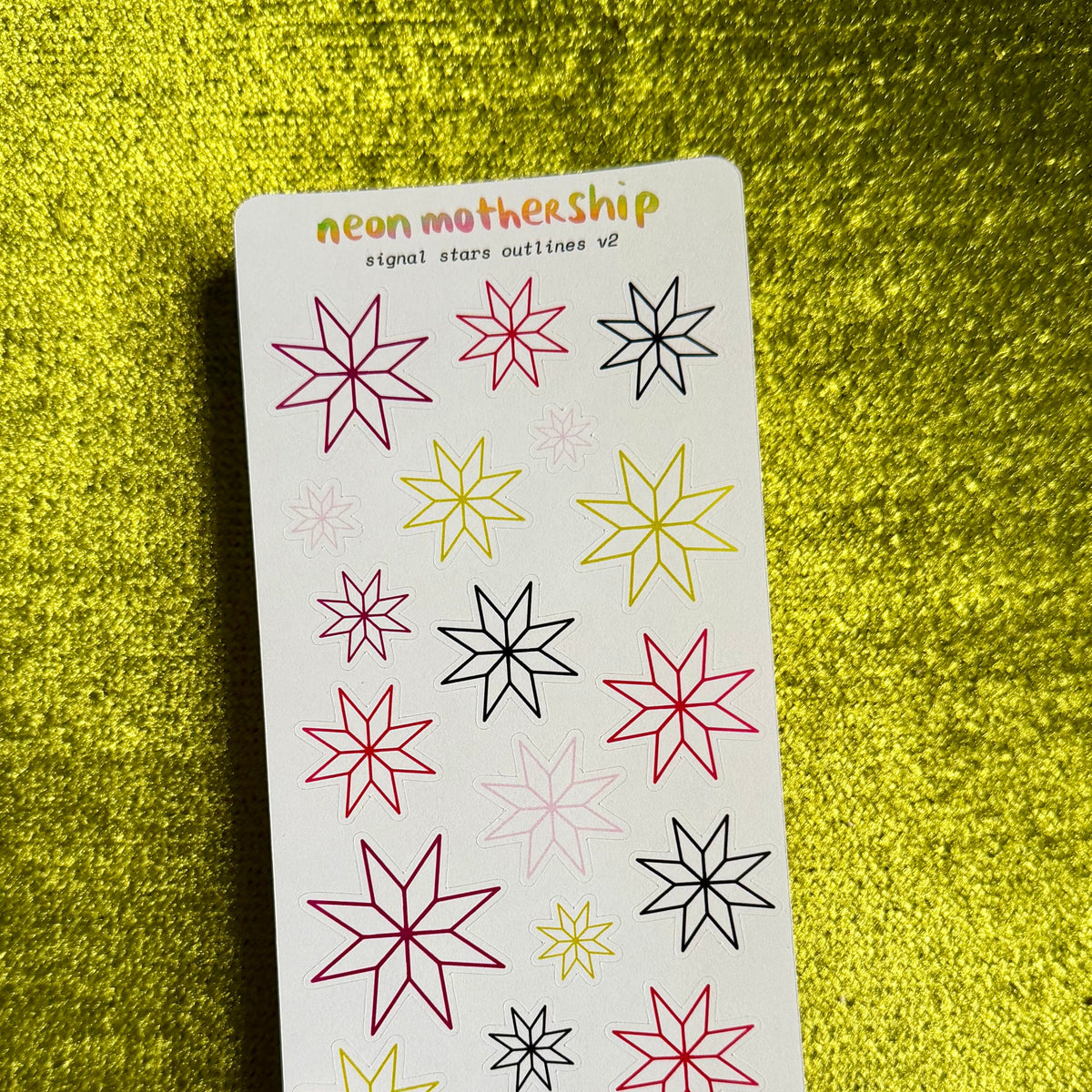A sticker sheet from the brand 'neon mothership' featuring various outlined star and snowflake designs in pink, yellow, and black on a white backing card, set against a textured yellow background.