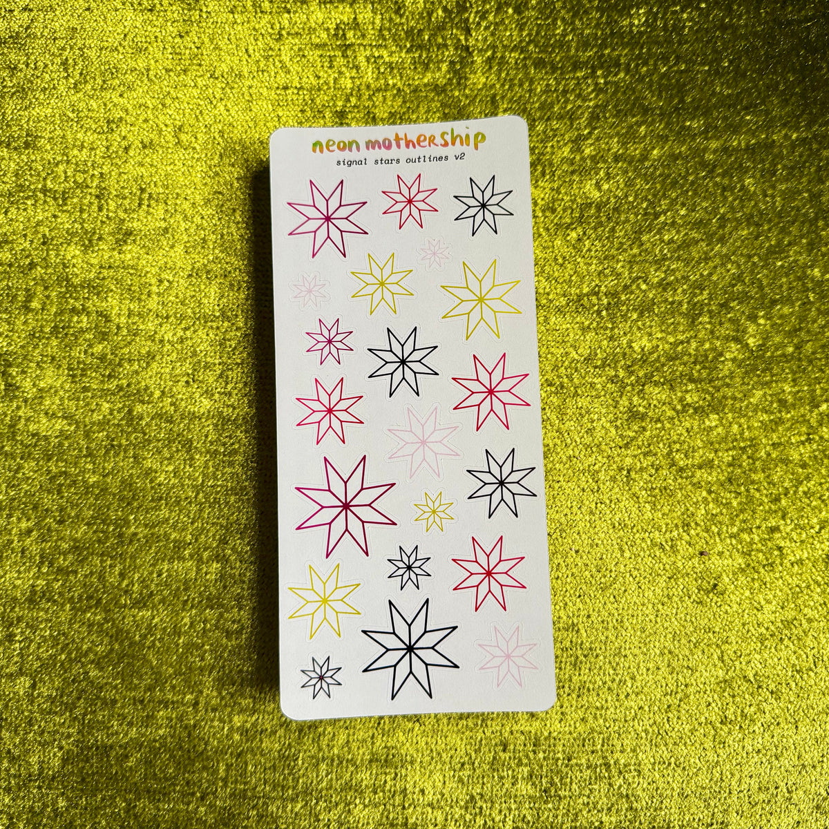 A rectangular white sticker sheet labeled 'neon mothership' featuring multiple neon-colored starburst designs in pink, yellow, and black, displayed on a textured bright yellow background.