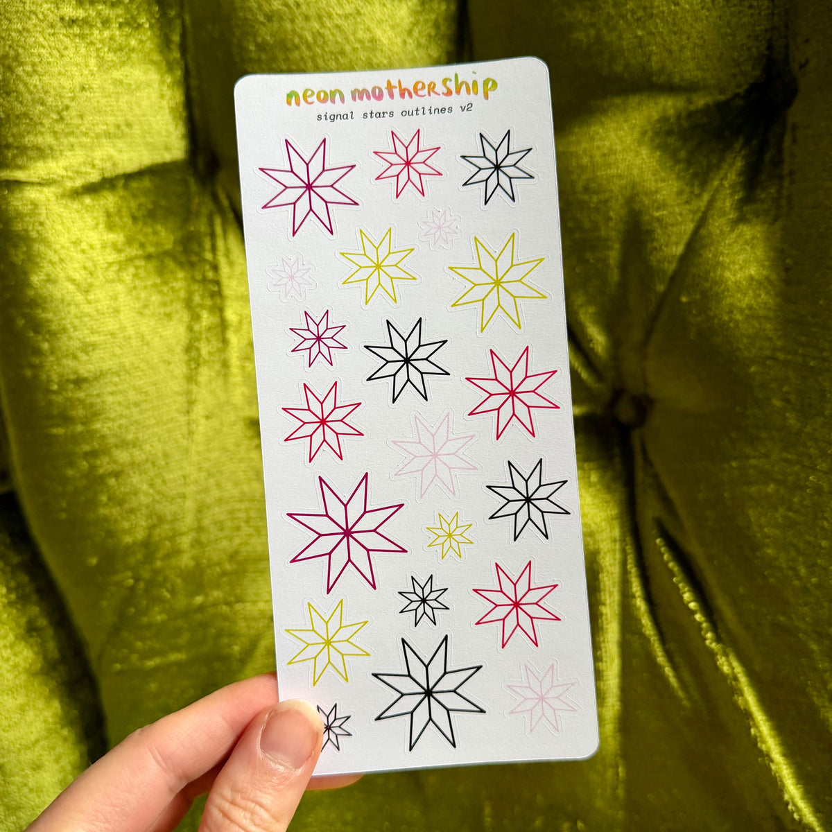 A hand holds a white sticker sheet featuring colorful neon star designs, set against a shiny olive green velvet background.