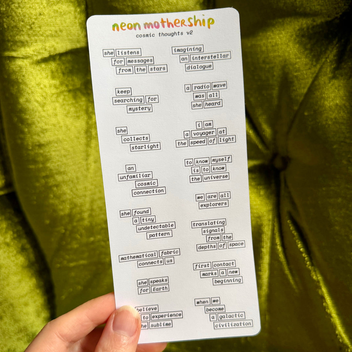 A hand holds a white sticker sheet titled 'neon mothership cosmic thoughts' against a shiny olive green fabric background. The sheet contains many small black text phrases and words.