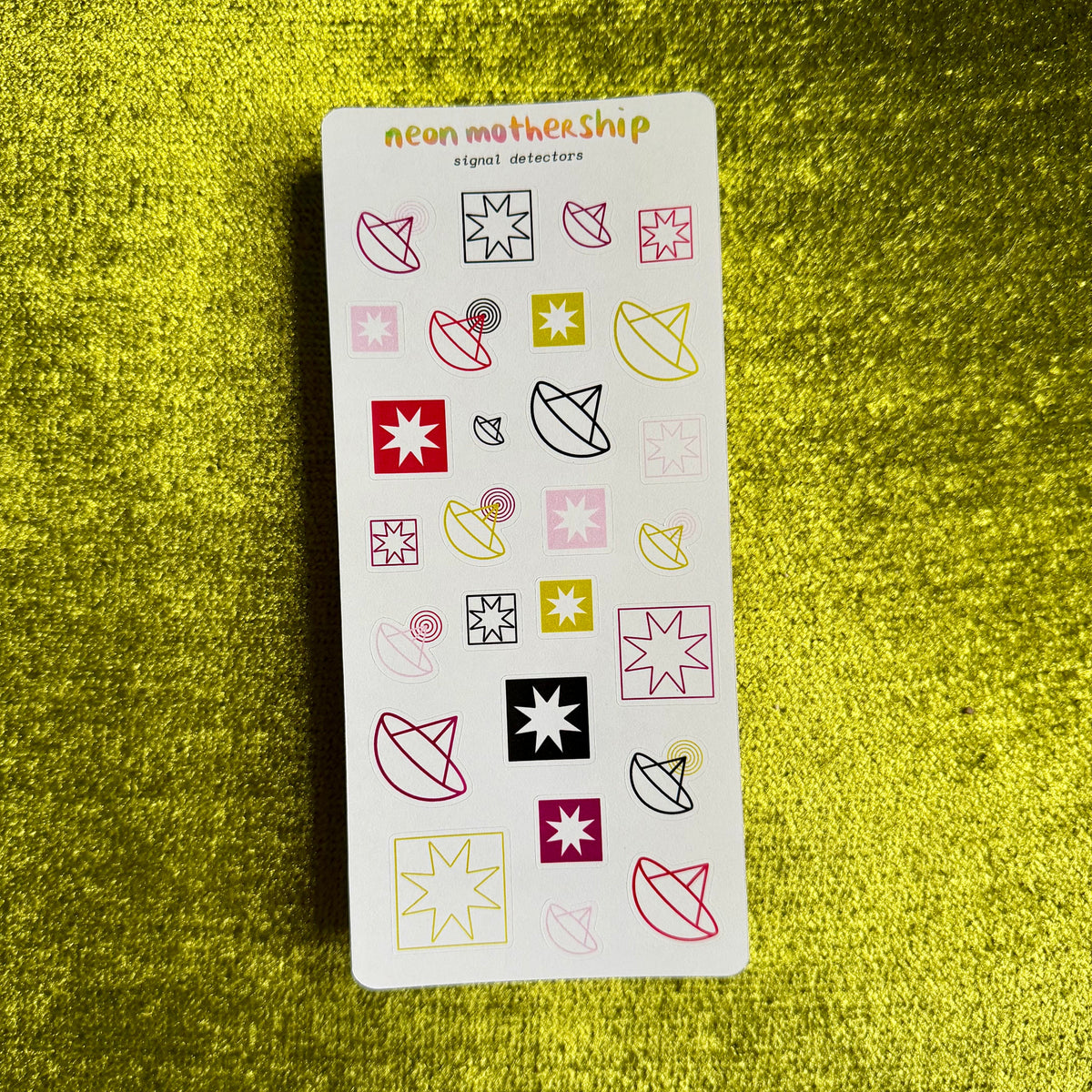 A white sticker sheet featuring various geometric and abstract shapes in neon pink, yellow, red, and black, displayed on a textured gold glitter background. The brand name 'neon motherShip' is printed at the top of the sheet.