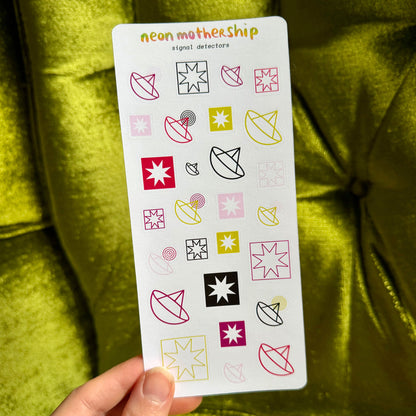 A hand holds a white sticker sheet labeled 'neon mothership signal detectors' against a shiny olive green fabric background. The sheet contains multiple colorful geometric and abstract neon-style icons in pink, yellow, black, and red.