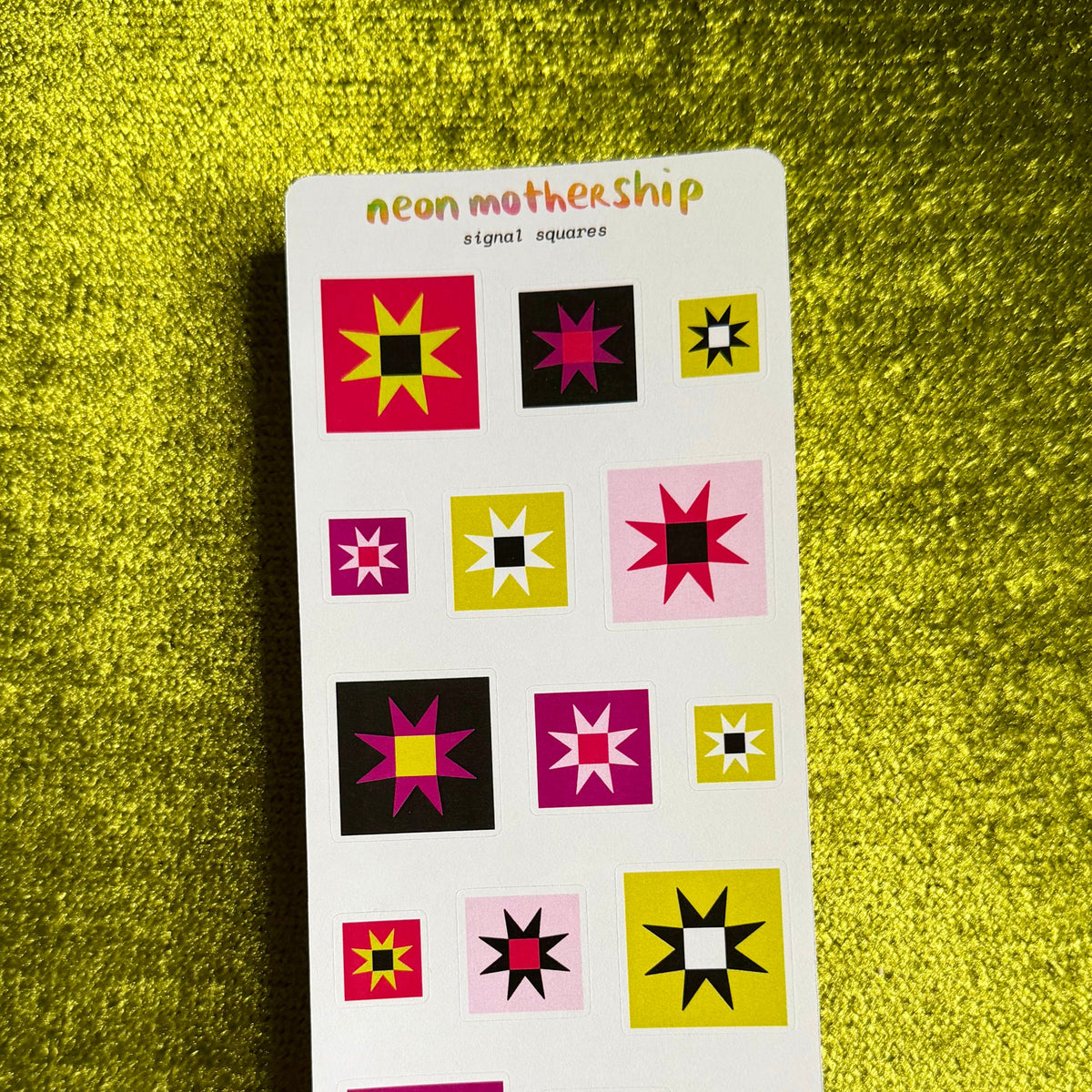 A vertical white sticker sheet labeled 'neon mothership signal squares' featuring multiple colorful geometric starburst and square designs, placed on a textured bright yellow-green surface.