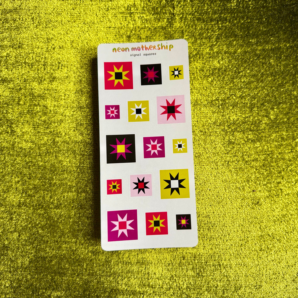 A sticker sheet titled 'neon mothership' featuring twelve colorful geometric starburst stickers in pink, yellow, black, and white, displayed on a textured, shiny gold green background.