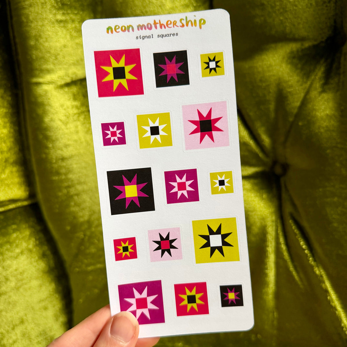A hand holds a white sticker sheet labeled 'neon mothership signal squares' featuring multiple colorful geometric starburst and square motifs, set against a bright yellow-green velvet chair background.