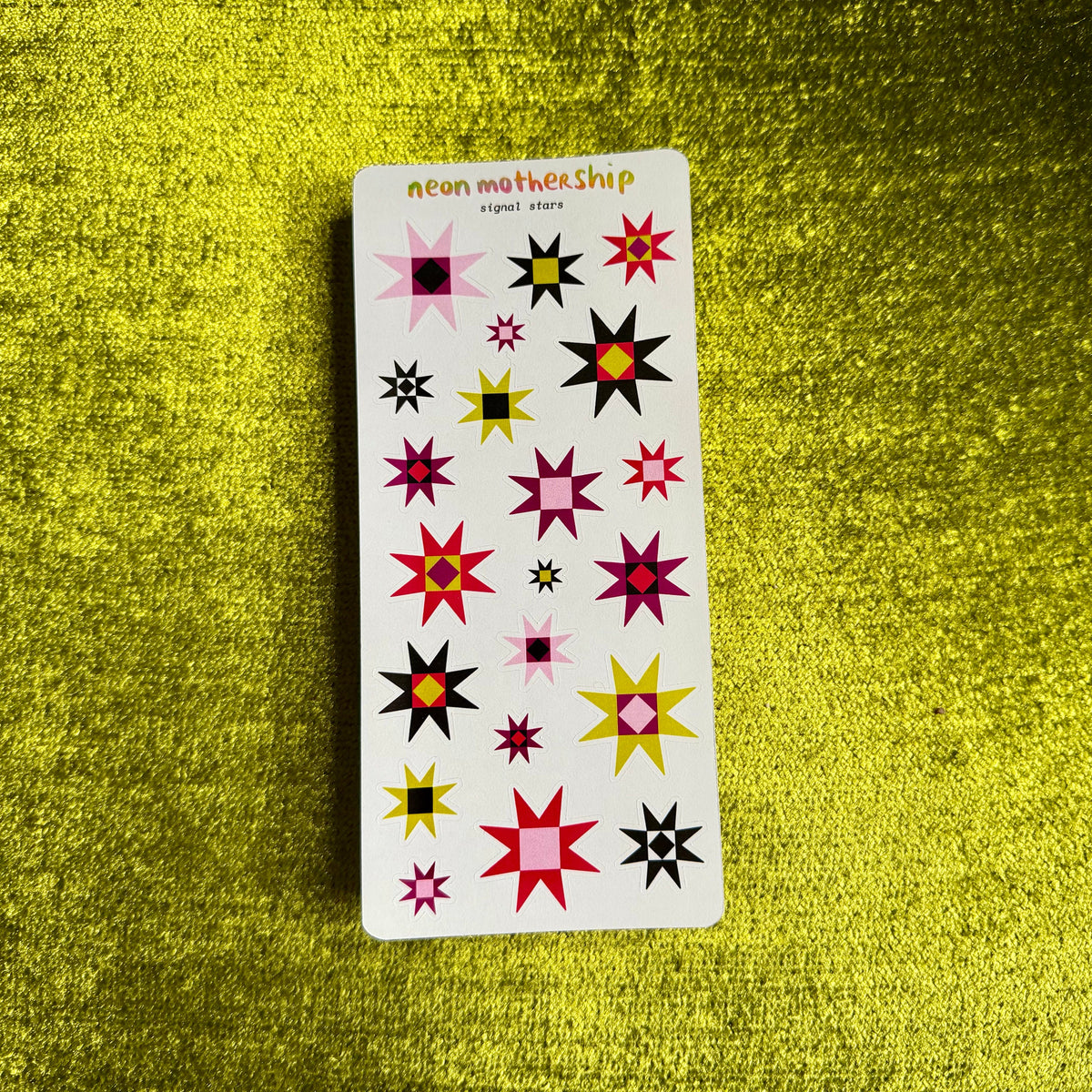 A white sticker sheet from the brand neon mothership, featuring various colorful starburst and geometric shapes, placed on a textured green background.