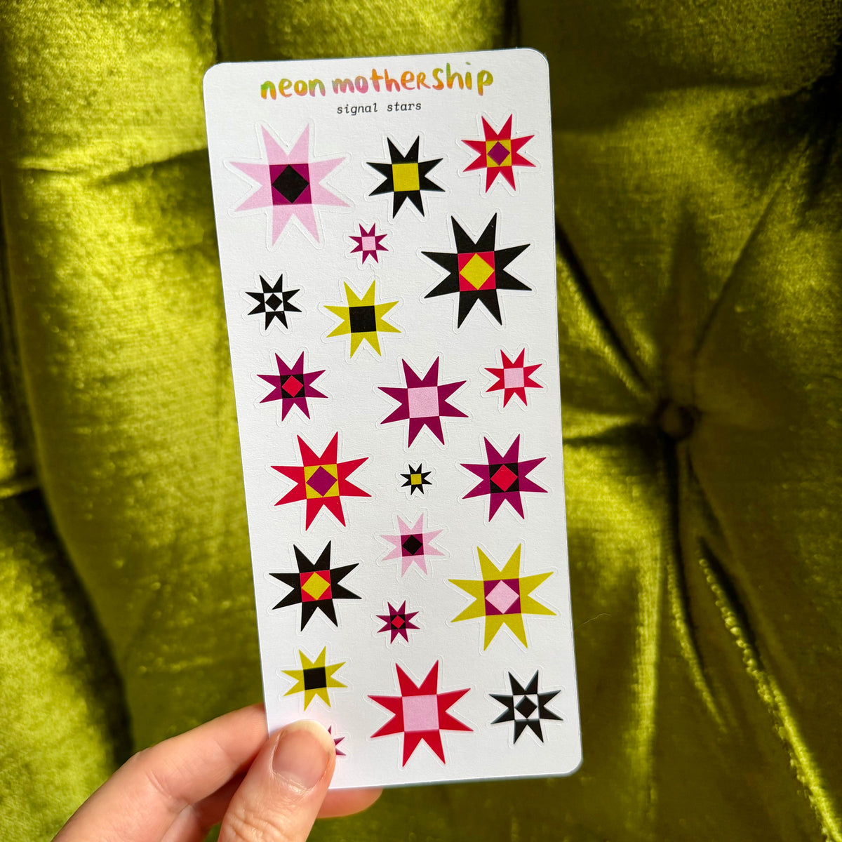 A hand holds a white card labeled 'neon mothership signal stars' against a shiny olive green velvet background. The card features multiple colorful starburst designs in pink, red, yellow, black, and white.