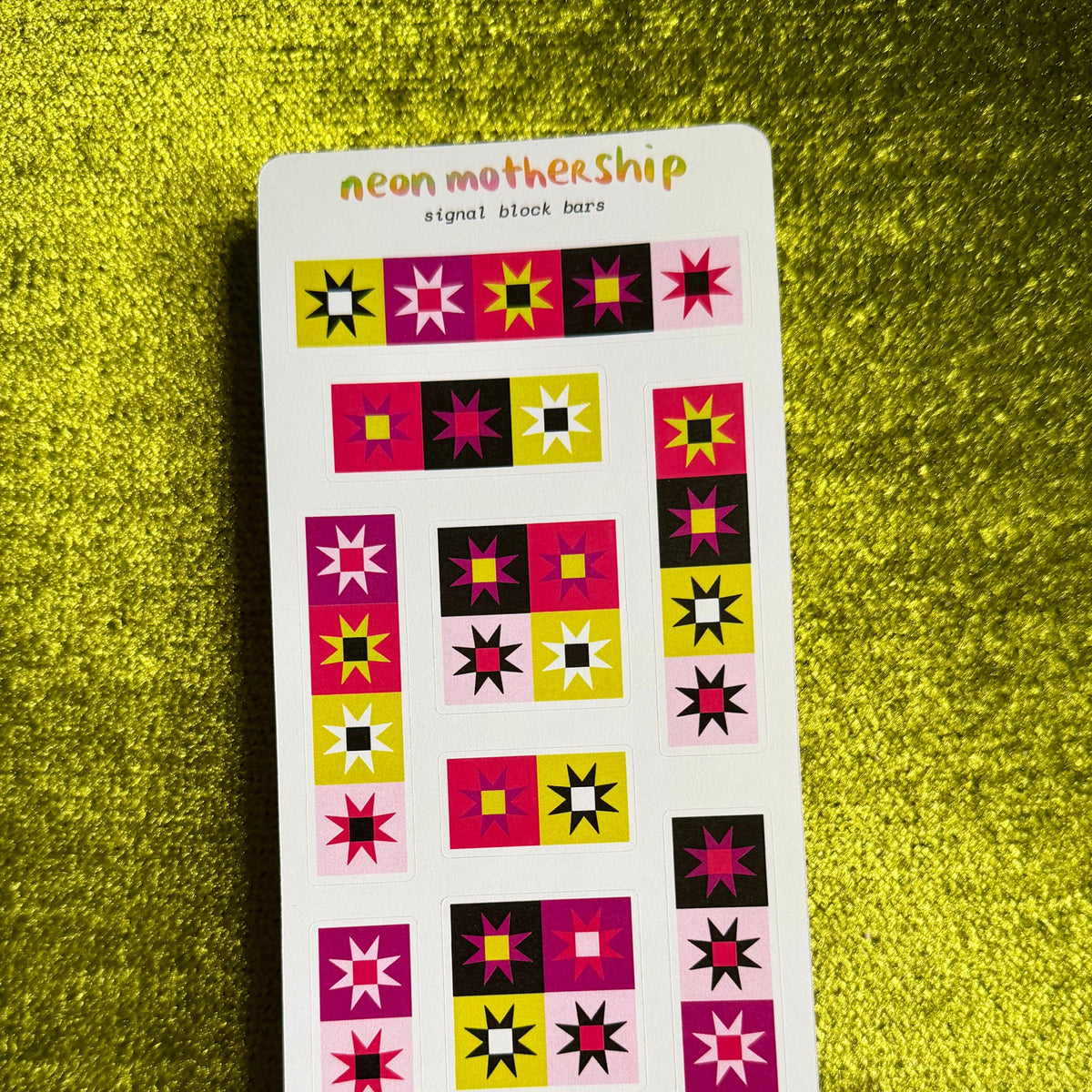 A sheet of decorative stickers with colorful geometric star and square patterns, labeled 'neon mothership' at the top, placed on a textured yellow-green surface.