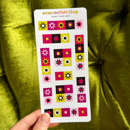 A hand holds a white sticker sheet labeled 'neon motherShip' against a shiny olive green velvet background. The sheet contains multiple colorful geometric starburst and square patch designs in pink, yellow, black, and white.