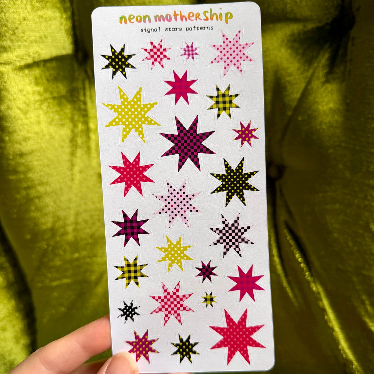 A hand holds a white sticker sheet featuring colorful neon starburst patterns under the text 'neon mothership signal star patterns'. The background is a textured olive green fabric.