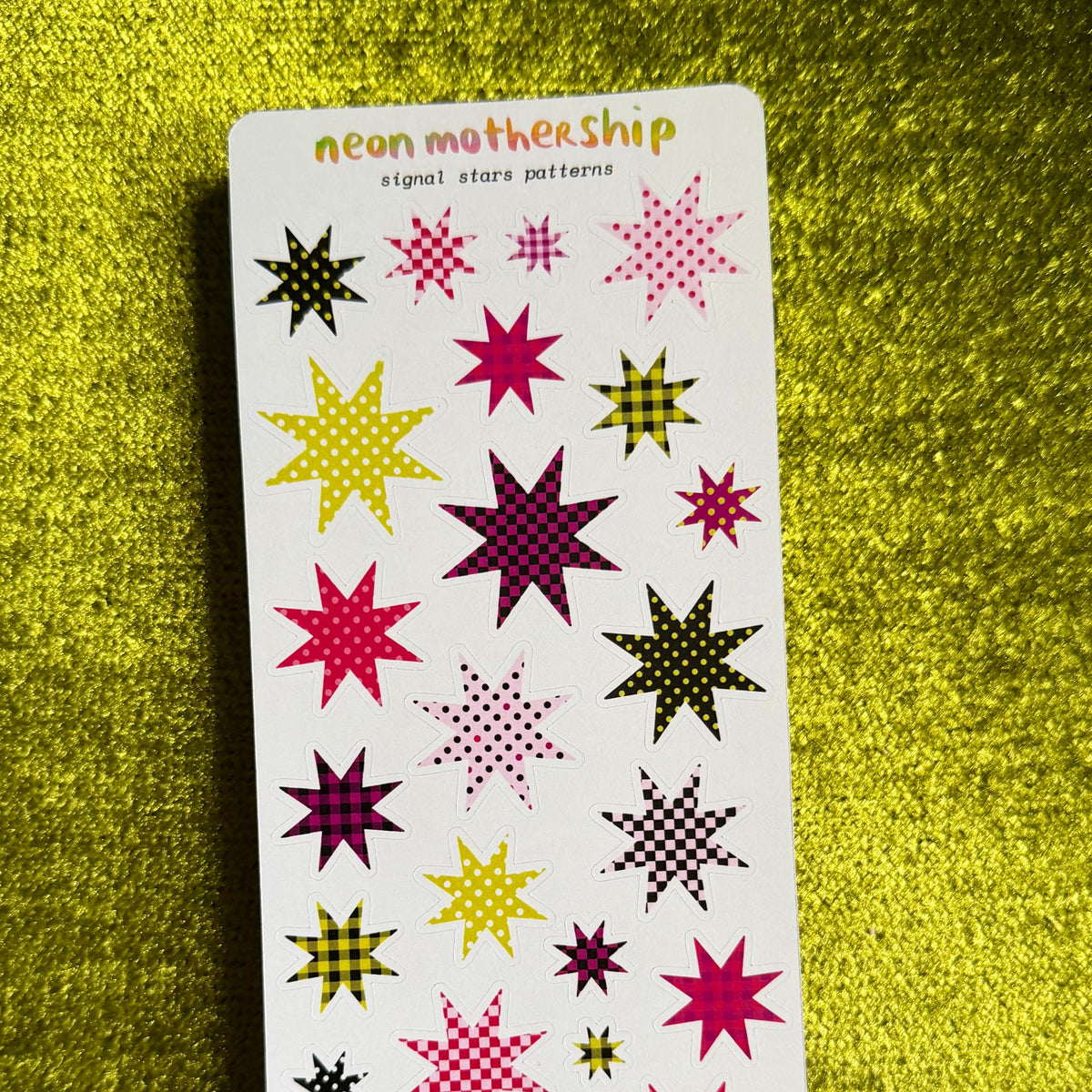 A sheet of neon mothership star patterns on a white backing card, displayed on a textured yellow-green background. The stars feature various neon colors including pink, purple, yellow, and green.