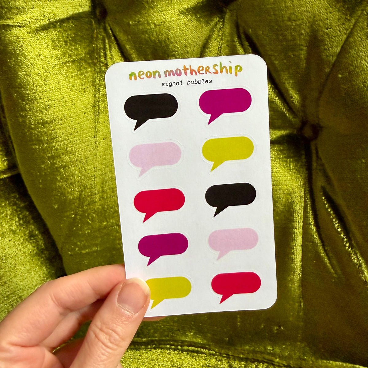 A hand holds a white sticker sheet labeled 'neon mothership signal bubbles' against a shiny olive green fabric background. The sheet contains colorful speech-bubble-shaped stickers in black, magenta, pink, yellow, and red.