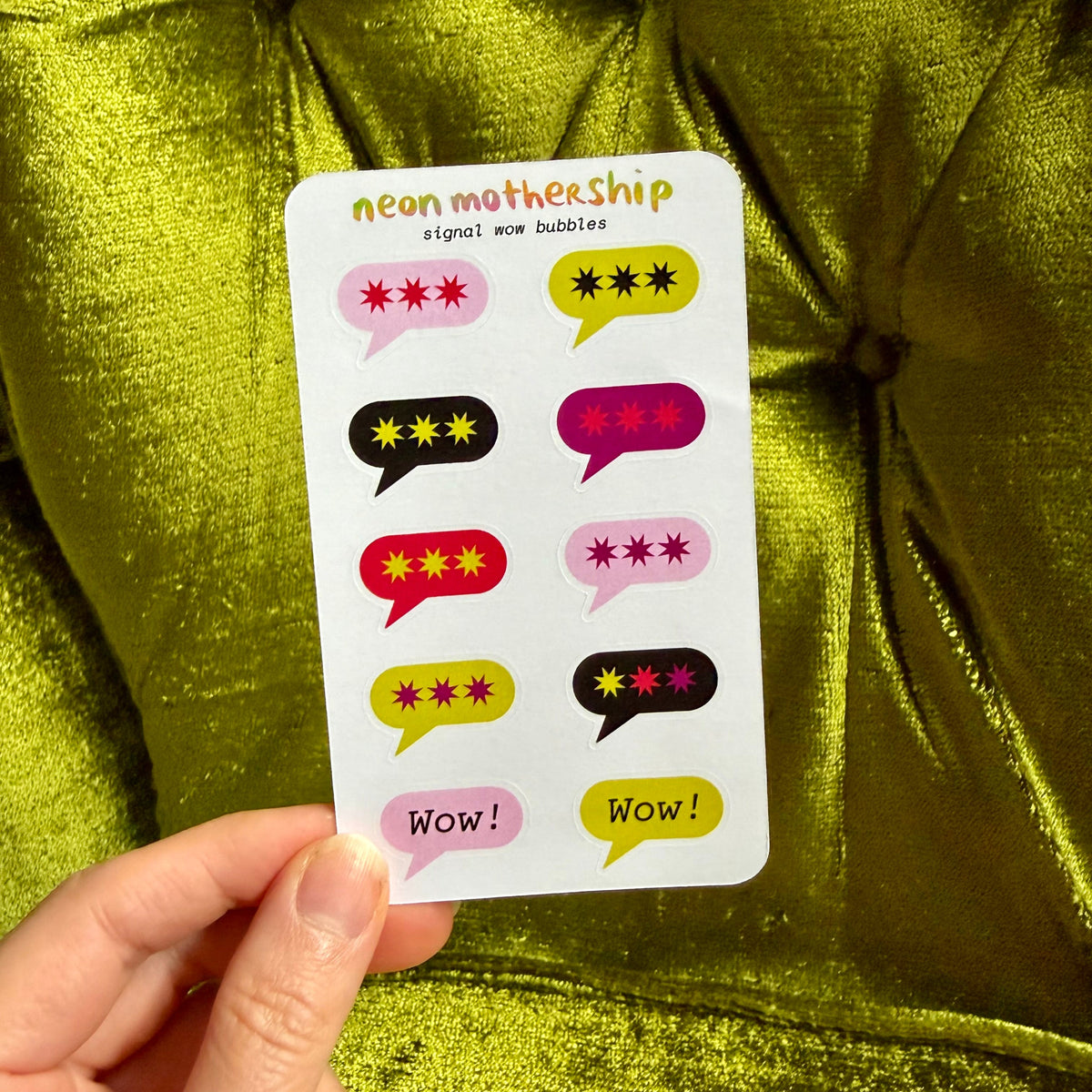 A hand holds a white sticker sheet featuring colorful speech bubble graphics with star ratings and the text 'neon mothership' and 'signal wow bubbles', set against a shiny olive green fabric background.