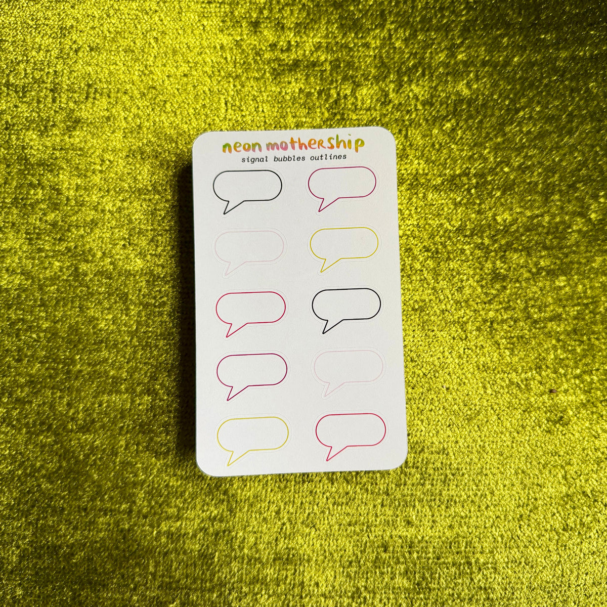A white sticker sheet labeled 'neon mothership' with multiple speech bubble outline stickers in neon pink, yellow, and black, placed on a textured yellow-green fabric background.