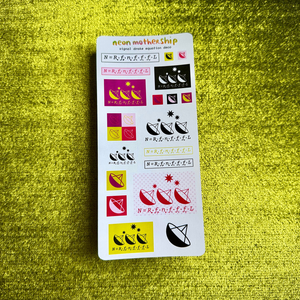 A white sticker sheet from the brand neon mothership, featuring various geometric and celestial designs in black, pink, yellow, and red, laid flat on a textured yellow-green fabric background.
