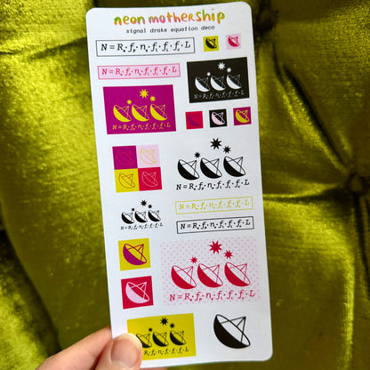 A hand holds a white sticker sheet featuring neon pink, yellow, and black mathematical equation and cat-themed signal decode decals, set against a shiny olive green fabric background.