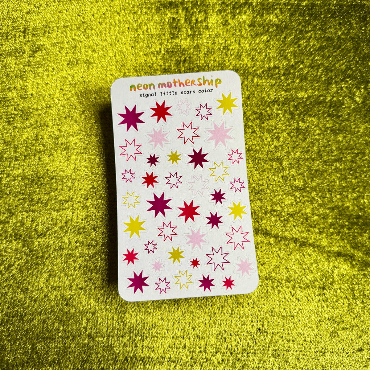 A rectangular sticker sheet from the brand neon mothership, featuring colorful star-shaped stickers in pink, yellow, and purple on a white background, placed on a textured yellow surface.