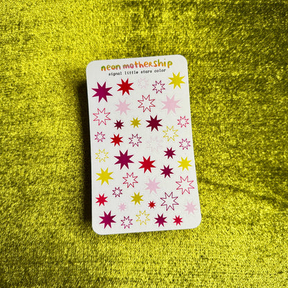 A rectangular sticker sheet from the brand neon mothership, featuring colorful star-shaped stickers in pink, yellow, and purple on a white background, placed on a textured yellow surface.