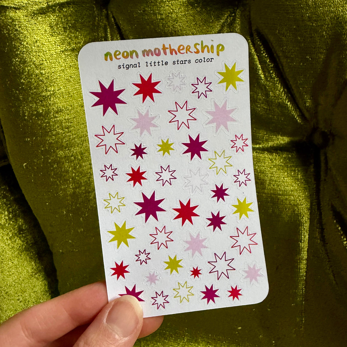 A hand holds a sticker sheet labeled 'neon mothership signal little stars color' featuring various neon star designs in pink, red, yellow, and purple, set against a shiny olive green fabric background.