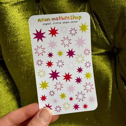 A hand holds a sticker sheet labeled 'neon mothership signal little stars color' featuring various neon star designs in pink, red, yellow, and purple, set against a shiny olive green fabric background.