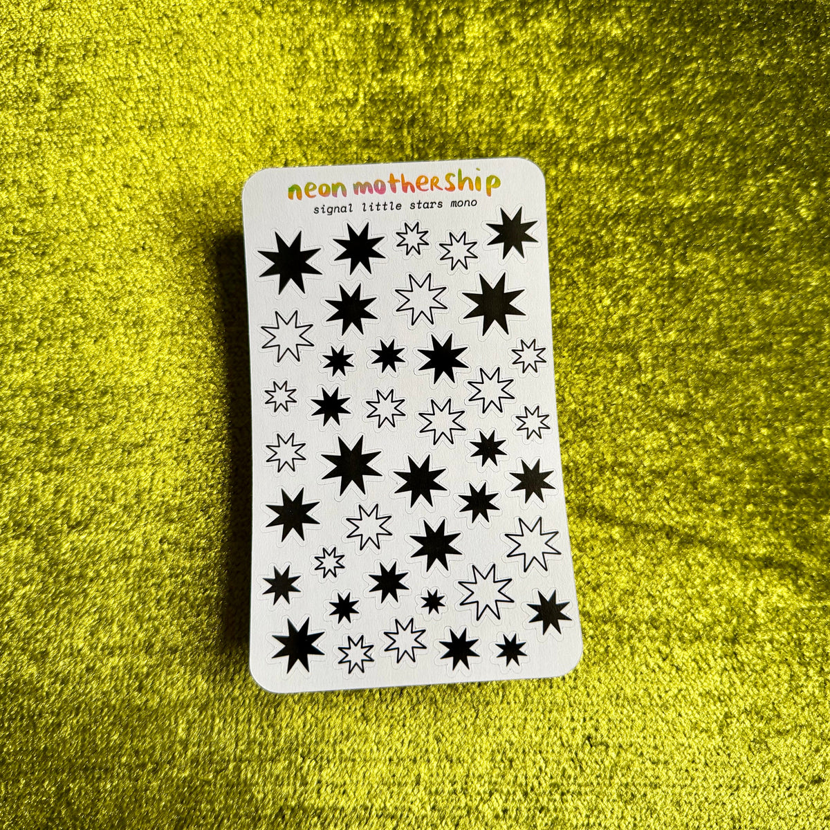 A white sticker sheet with black neon star designs and the text 'neon mothership signal: little star mono' lies on a textured bright yellow surface.