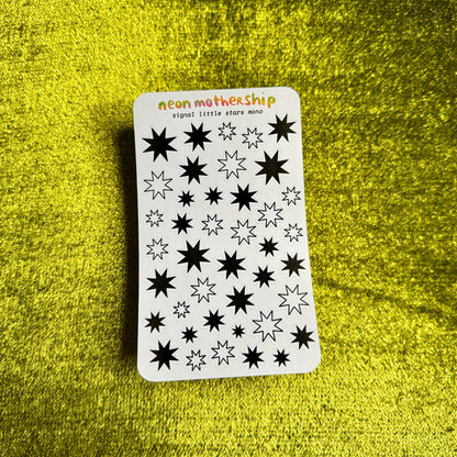 A white sticker sheet with black neon star designs and the text 'neon mothership signal: little star mono' lies on a textured bright yellow surface.