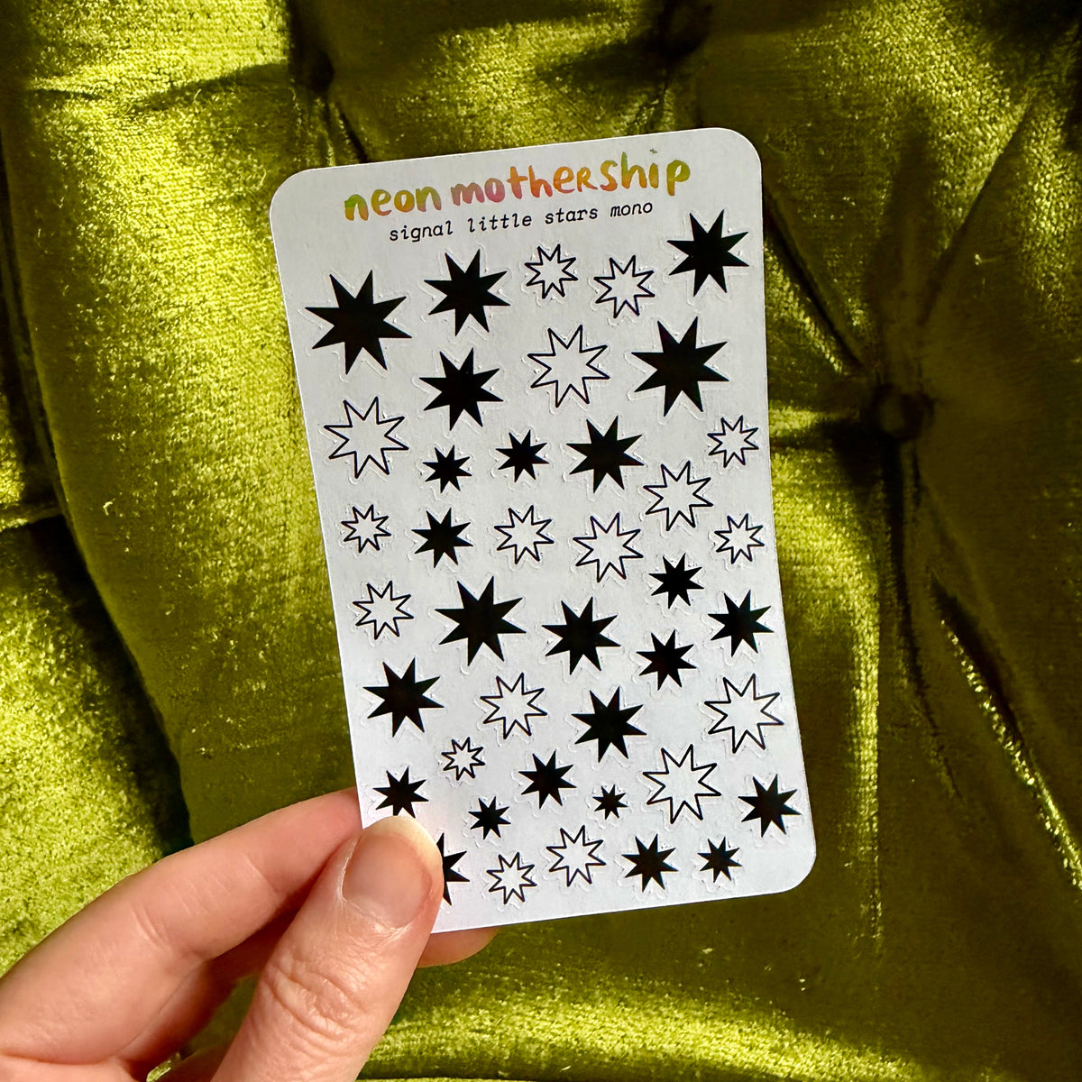 A hand holds a white sticker sheet featuring various black and white neon star designs, with the text 'neon mothership signal little stars mono!' printed at the top, set against a shiny olive green fabric background.