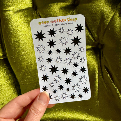 A hand holds a white sticker sheet featuring various black and white neon star designs, with the text 'neon mothership signal little stars mono!' printed at the top, set against a shiny olive green fabric background.